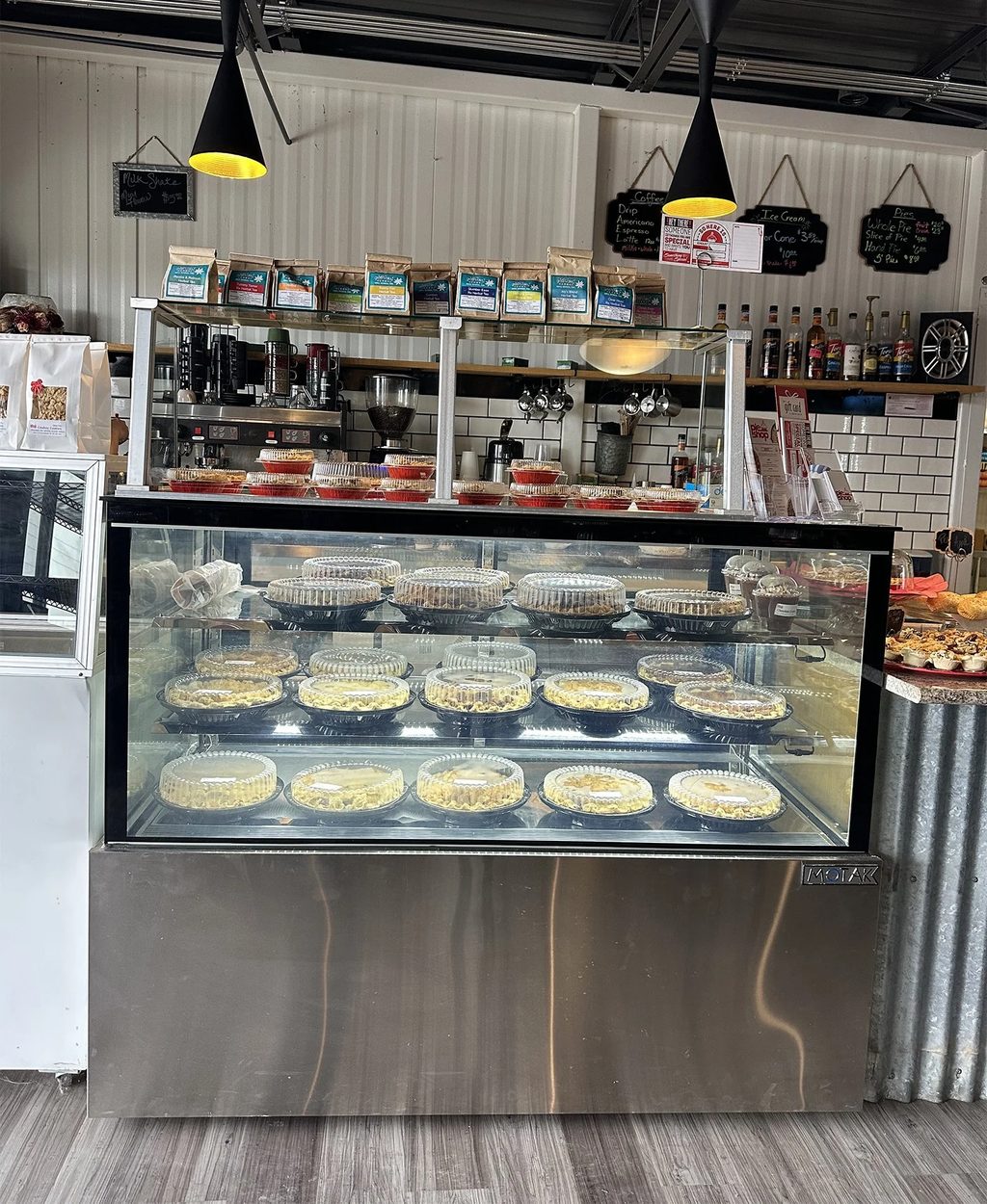 The pie shop's glass display case filled with covered whole pies, with a coffee counter, chalkboard menus and syrup bottles behind it.